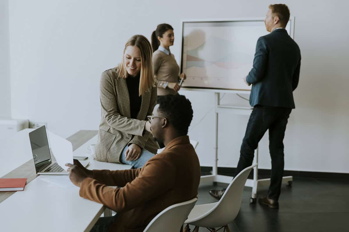 Four colleagues collaborate in an office, two at a desk with a laptop and two near a presentation board.