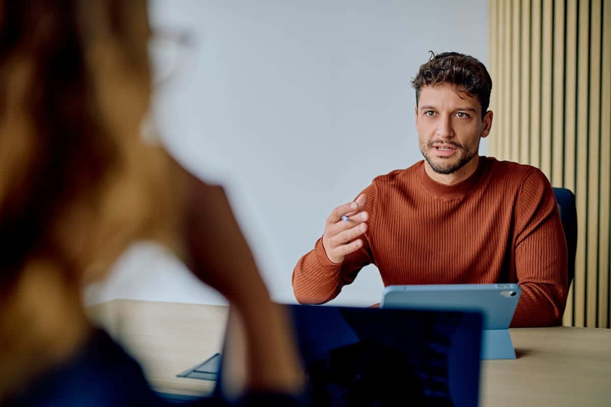 Man in brown sweater talking to a woman across a desk in an office setting, with a tablet in front of him.